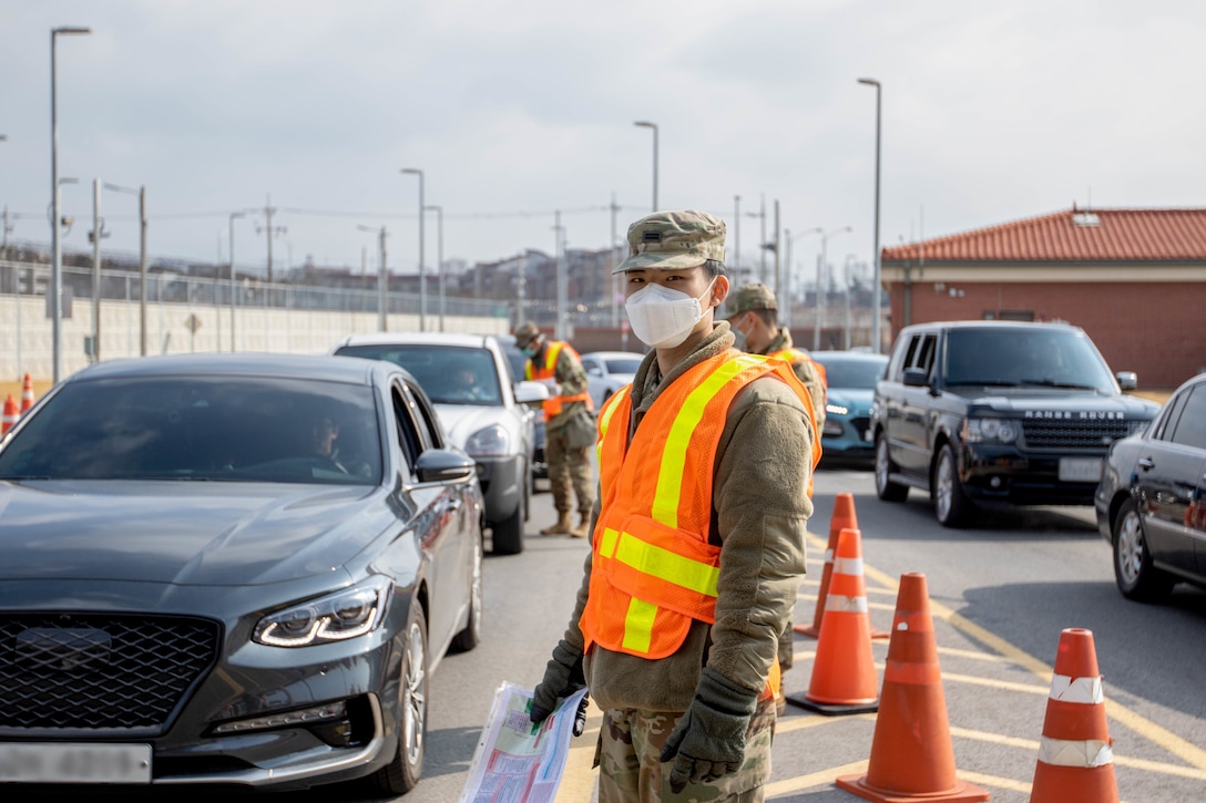 Soldier wearing gloves and a surgical mask stops vehicles at a checkpoint.