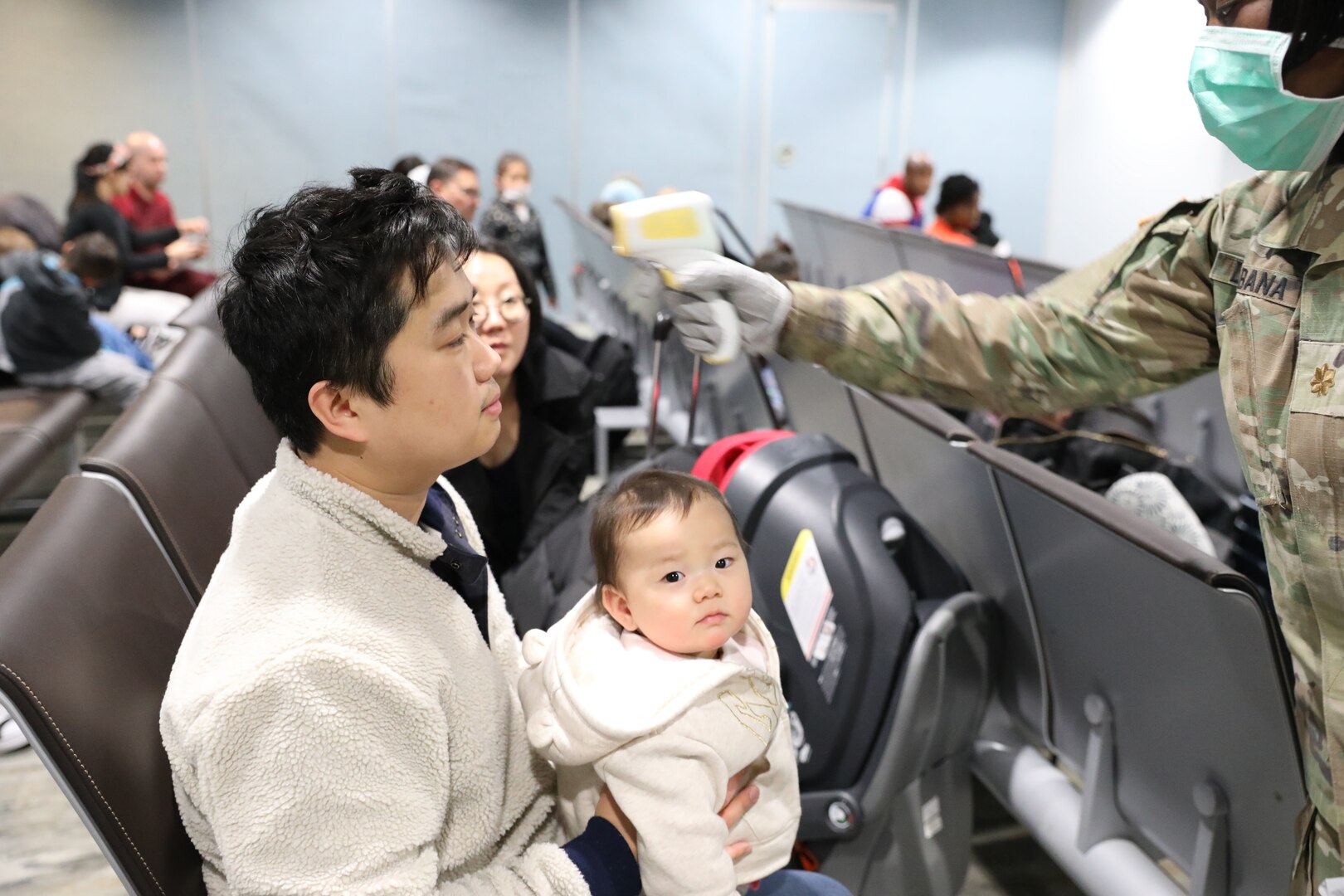 A military service member wearing a surgical mask holds a device to take the temperature of a family.