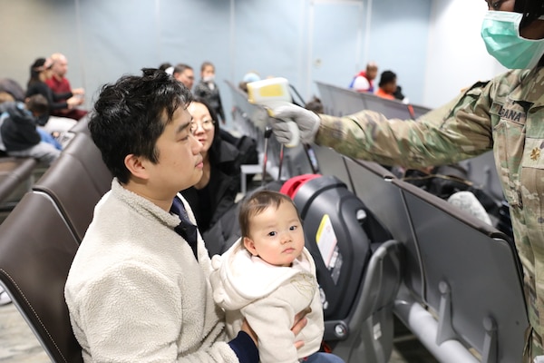 A military service member wearing a surgical mask holds a device to take the temperature of a family.