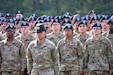 Men and women in green camouflage uniforms and black berets with blue flashes stand behind a woman in green camouflage uniform and green hat.