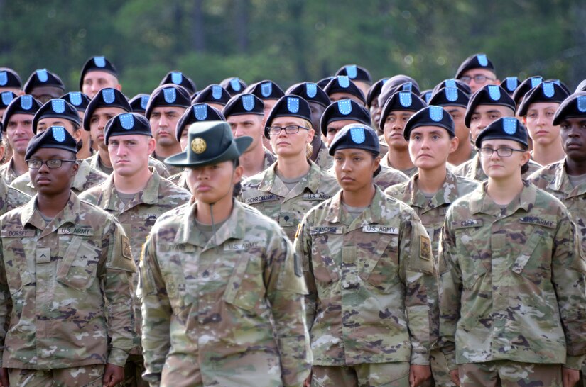 Men and women in green camouflage uniforms and black berets with blue flashes stand behind a woman in green camouflage uniform and green hat.