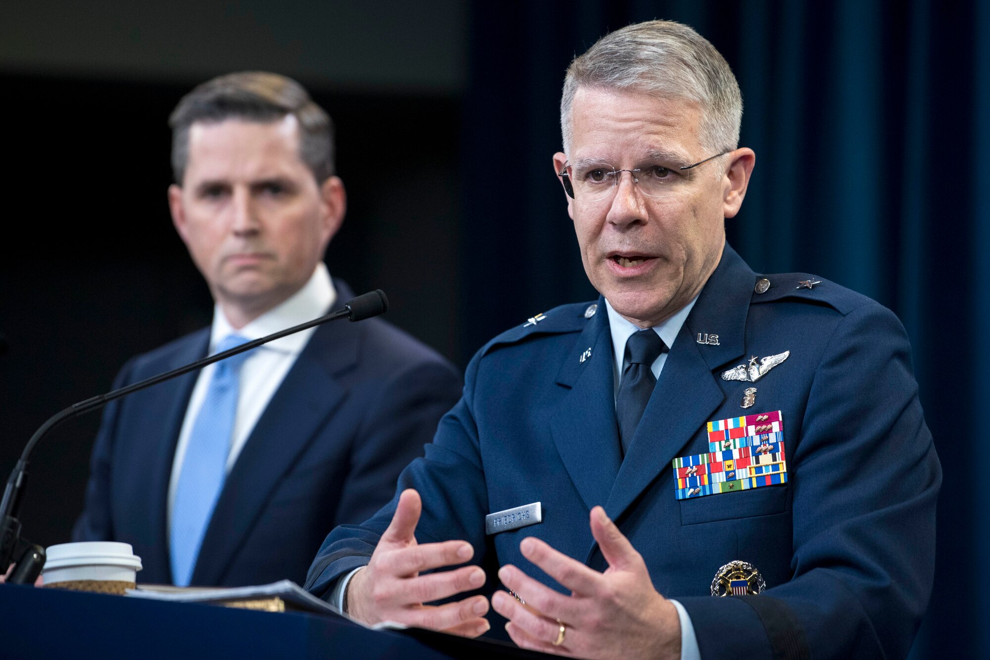 An airman speaks at a lectern while a civilian watches in the background.