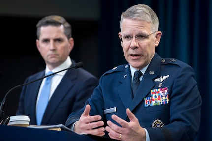 An airman speaks at a lectern while a civilian watches in the background.