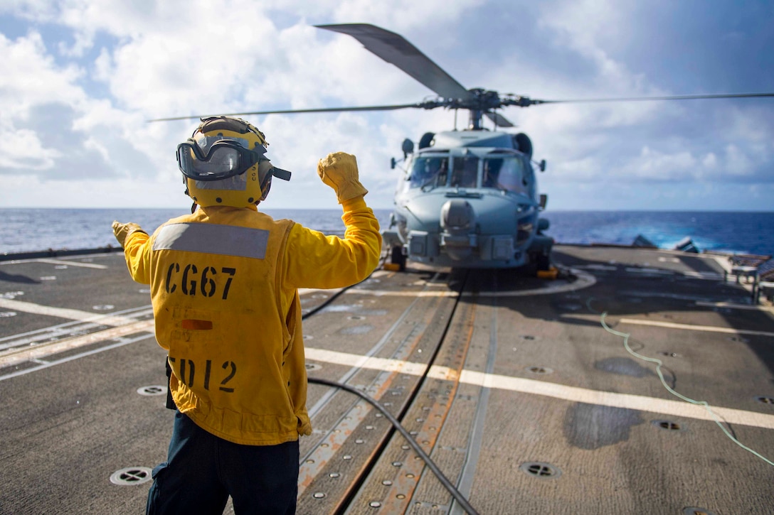 A sailor signals to a helicopter on the deck of a ship.