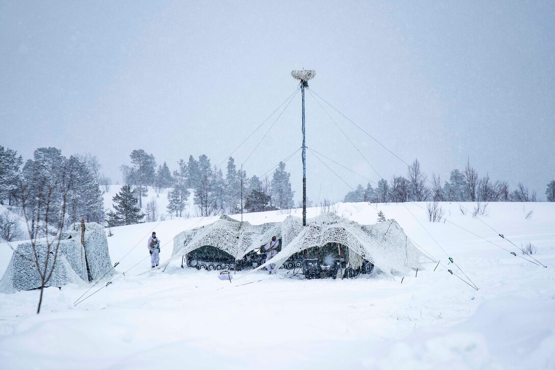 Marines stand next to military vehicles parked underneath netting in the snow.