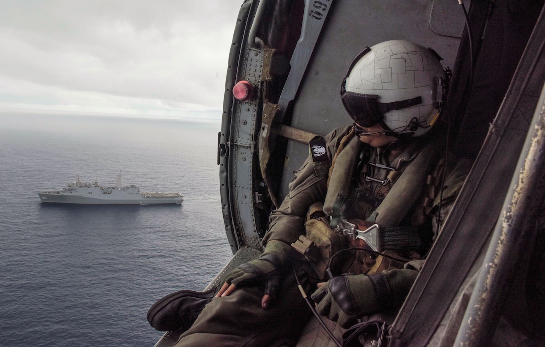 A sailor sits on the edge of a helicopter as a ship passes in the ocean.