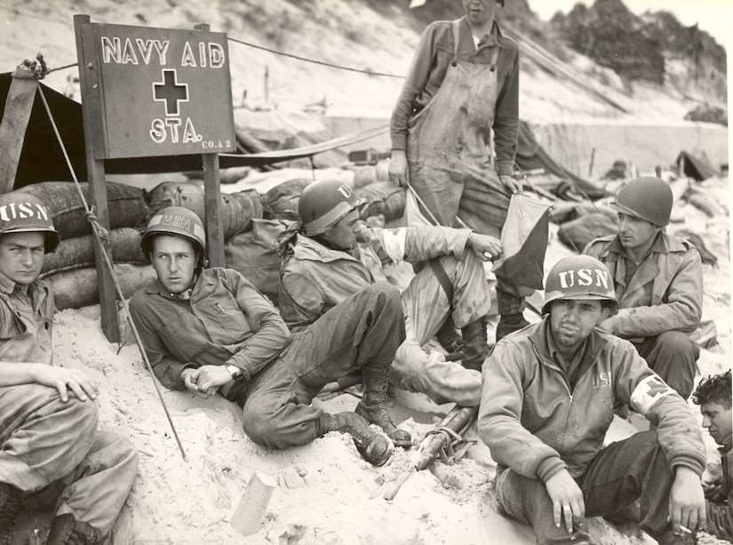 A group of servicemen sit on the sand; a sign in the background reads, “Navy Aid Sta.”