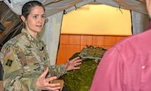 Woman in green camouflage uniform stands in a tent talking to person in red shirt.