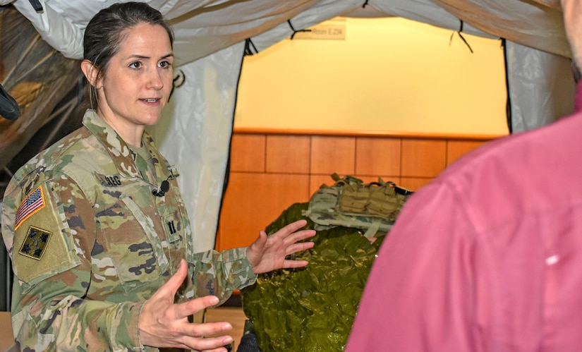 Woman in green camouflage uniform stands in a tent talking to person in red shirt.