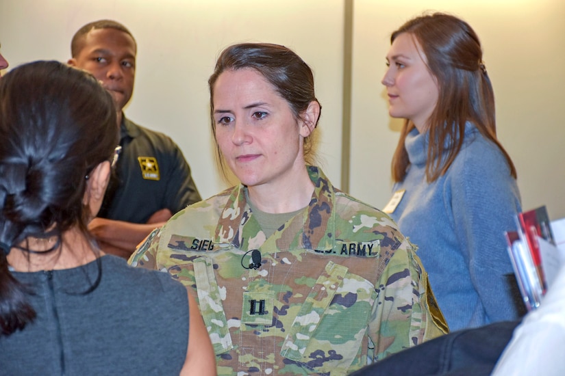 Woman in green camouflage uniforms stands in the middle of individuals in a room.