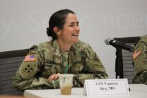 Female in green camouflage uniform sits in front of michrophone with arms crossed and smiles.