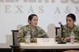 Two brown-haired women in green camouflage uniforms sit in black chairs behind a white table with a microphone.