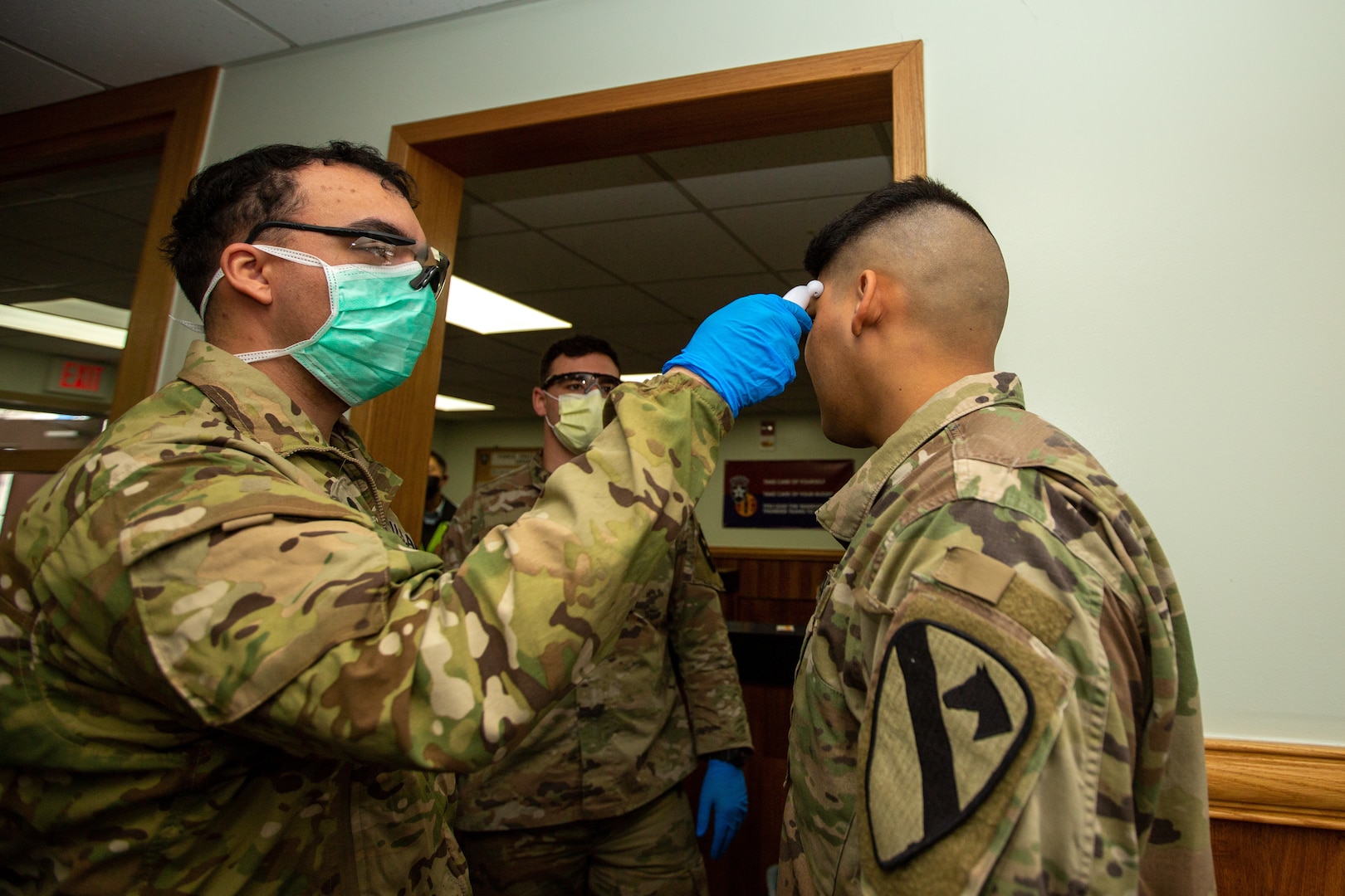 A uniformed soldier wears a face mask while taking the temperature of another soldier.