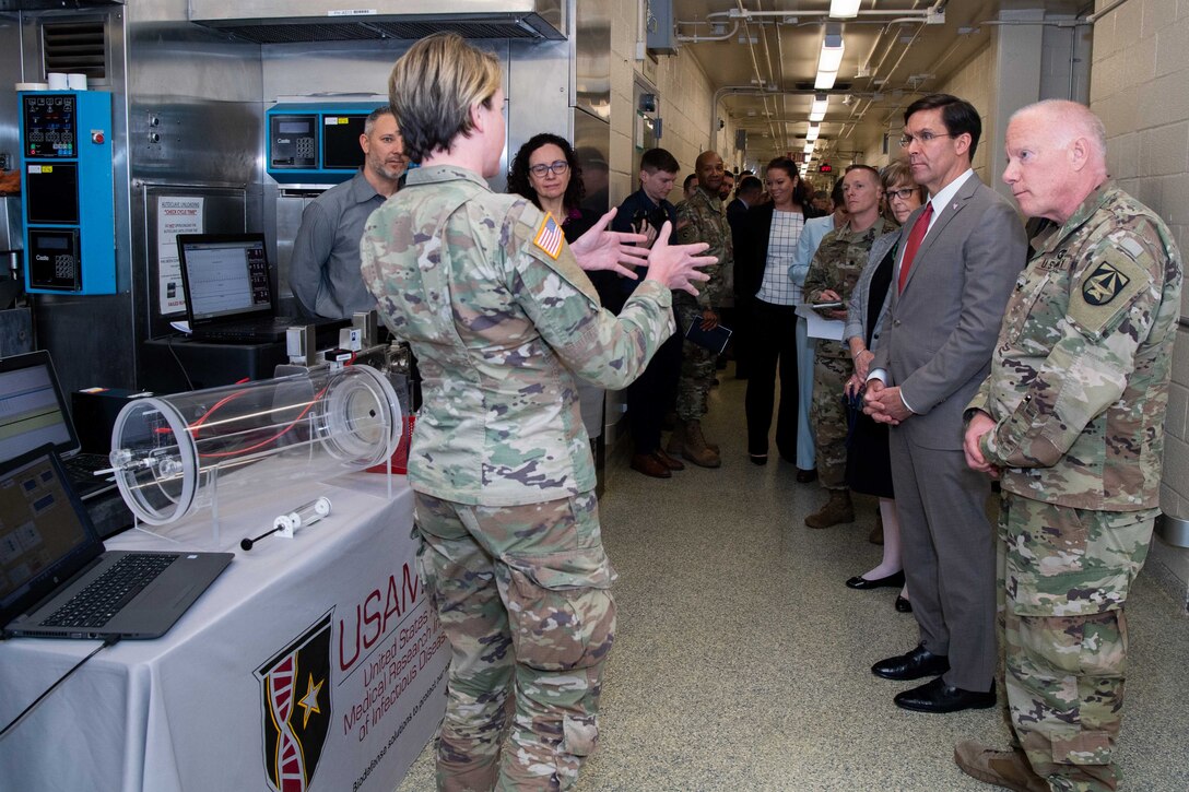 More than a dozen individuals stand facing a single female military member as she speaks about medical equipment on a table.