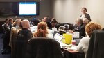 Group of people sit at a conference tables in front of a TV monitor while one woman stands and speaks.