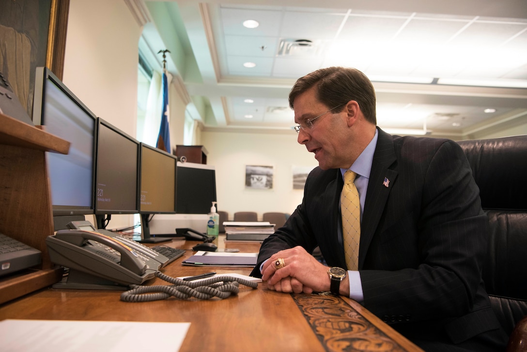 Defense Secretary Dr. Mark T. Esper sits and talks by a phone at a desk.