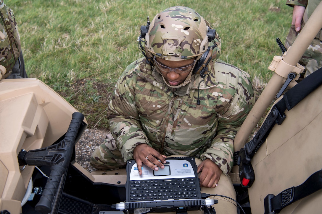 An airman, shown from overhead, sits on grass while using a keyboard on a portable device.