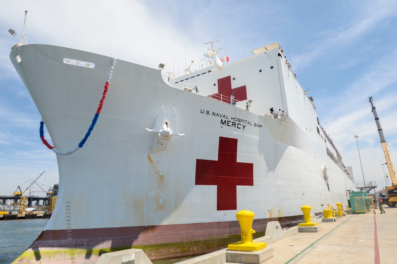 A large white ship is tied to a dock. The ship has the words “U.S. Naval Hospital Ship Mercy” on the side, along with a large red cross.