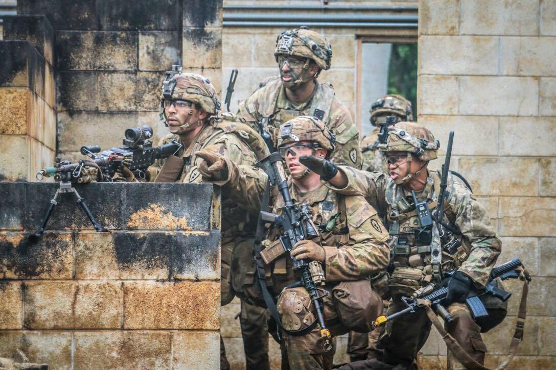 Soldiers gather behind a wall with weapons and look toward something in the distance.