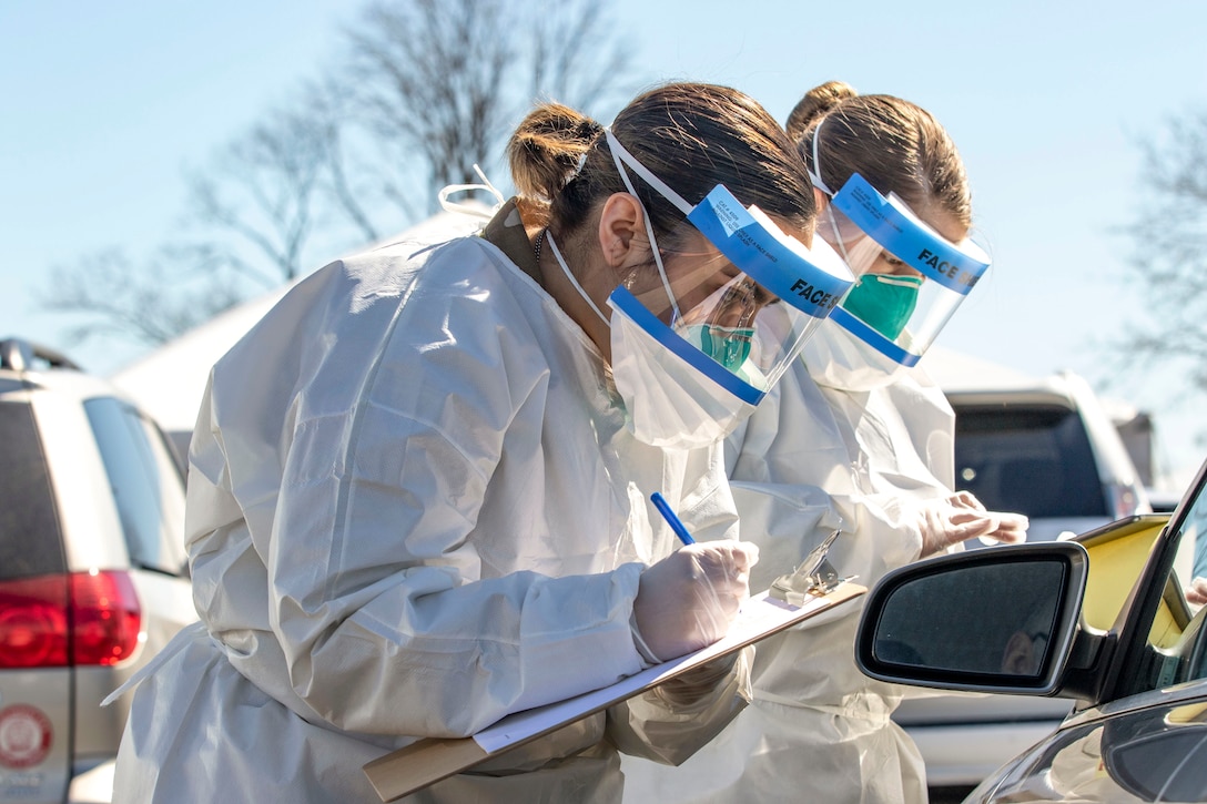 Two National Guardsmen wearing protective masks, gloves and smocks lean in toward the driver's side of a car and take notes.