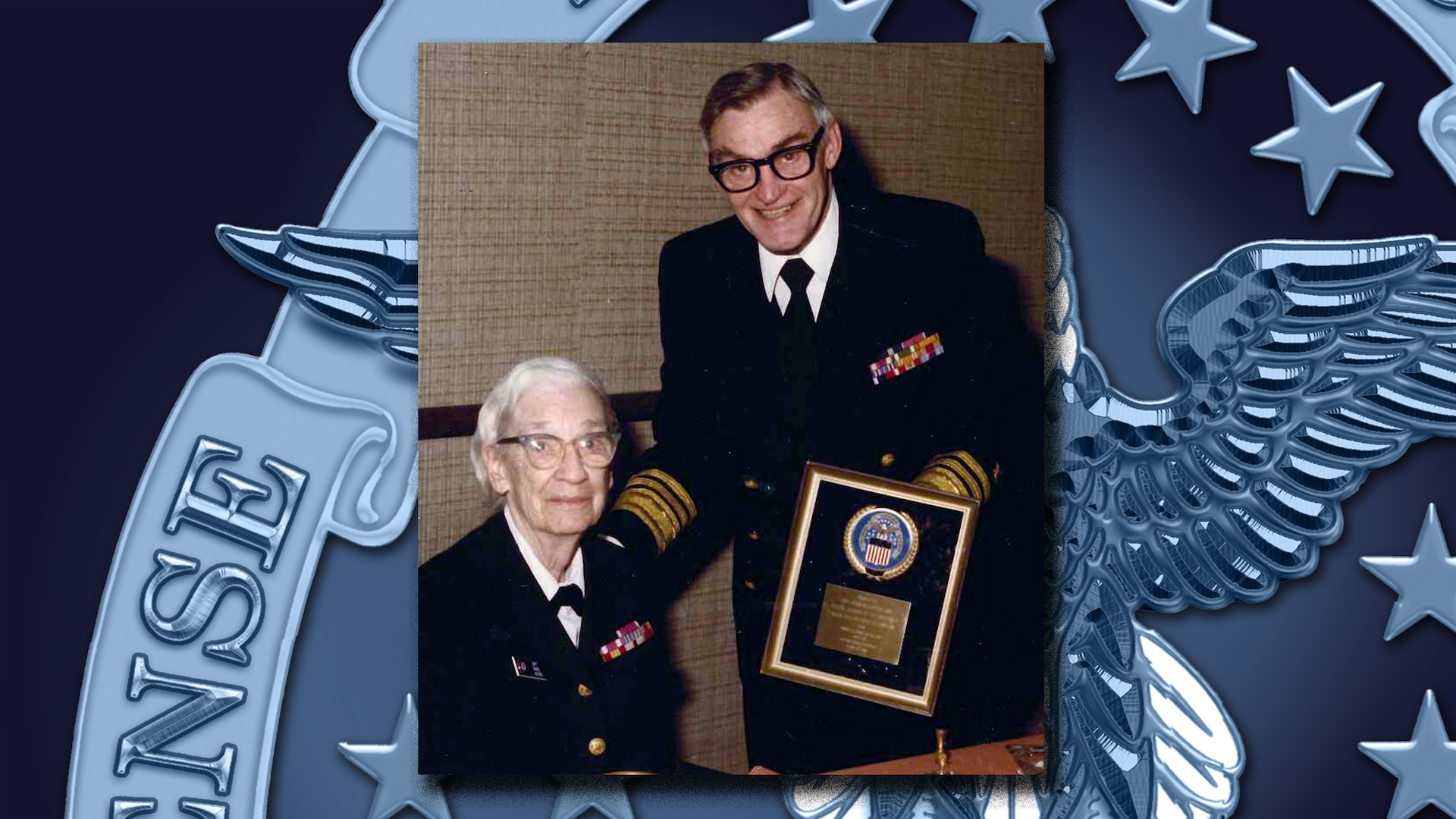 Man in dress Navy uniform holds a plaque he's presenting to a woman in a Navy dress uniform.
