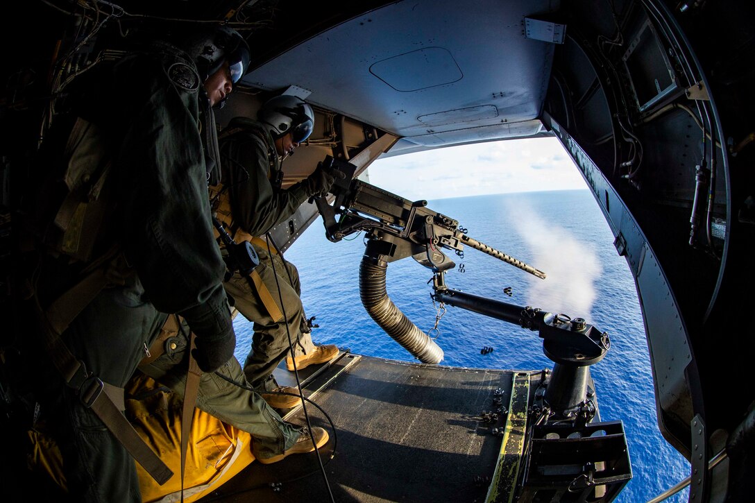 A Marine fires a machine gun out the door of a helicopter as another Marine stands behind.