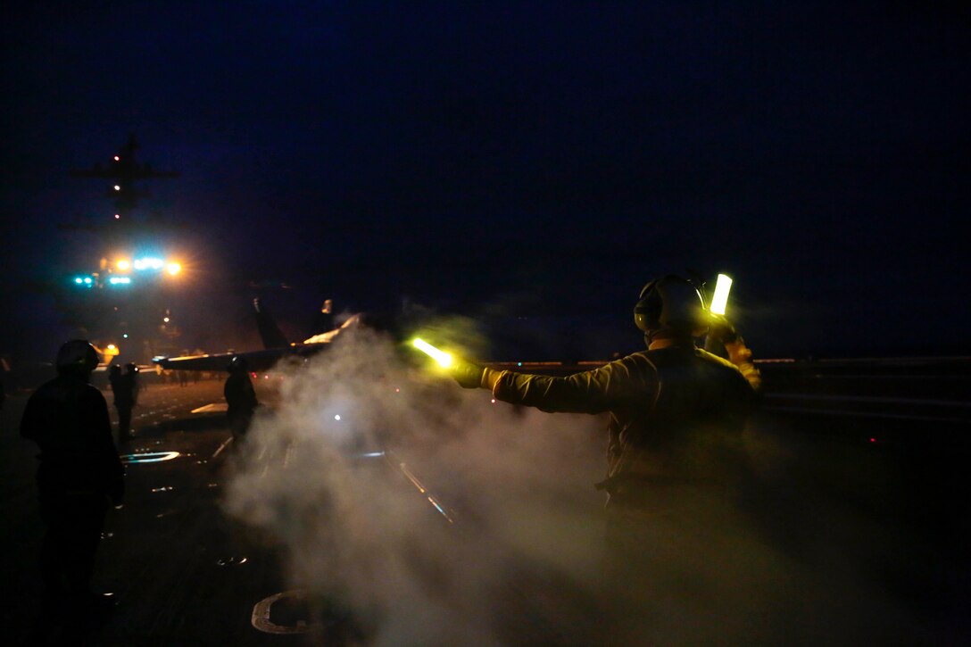 A sailor holds glow sticks in both hands to signal toward an aircraft at night.