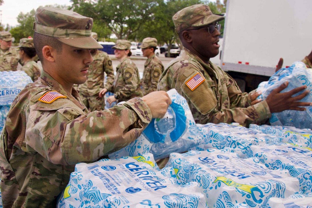 Two soldiers unload water cases from a vehicle.