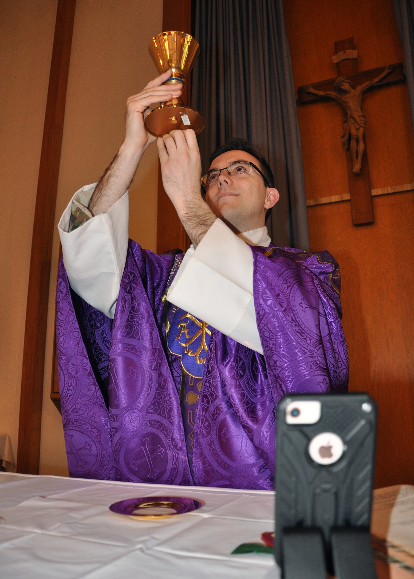 priest at altar holding up raised chalice for blessing with cell phone in foreground recording the mass for virtual reality.