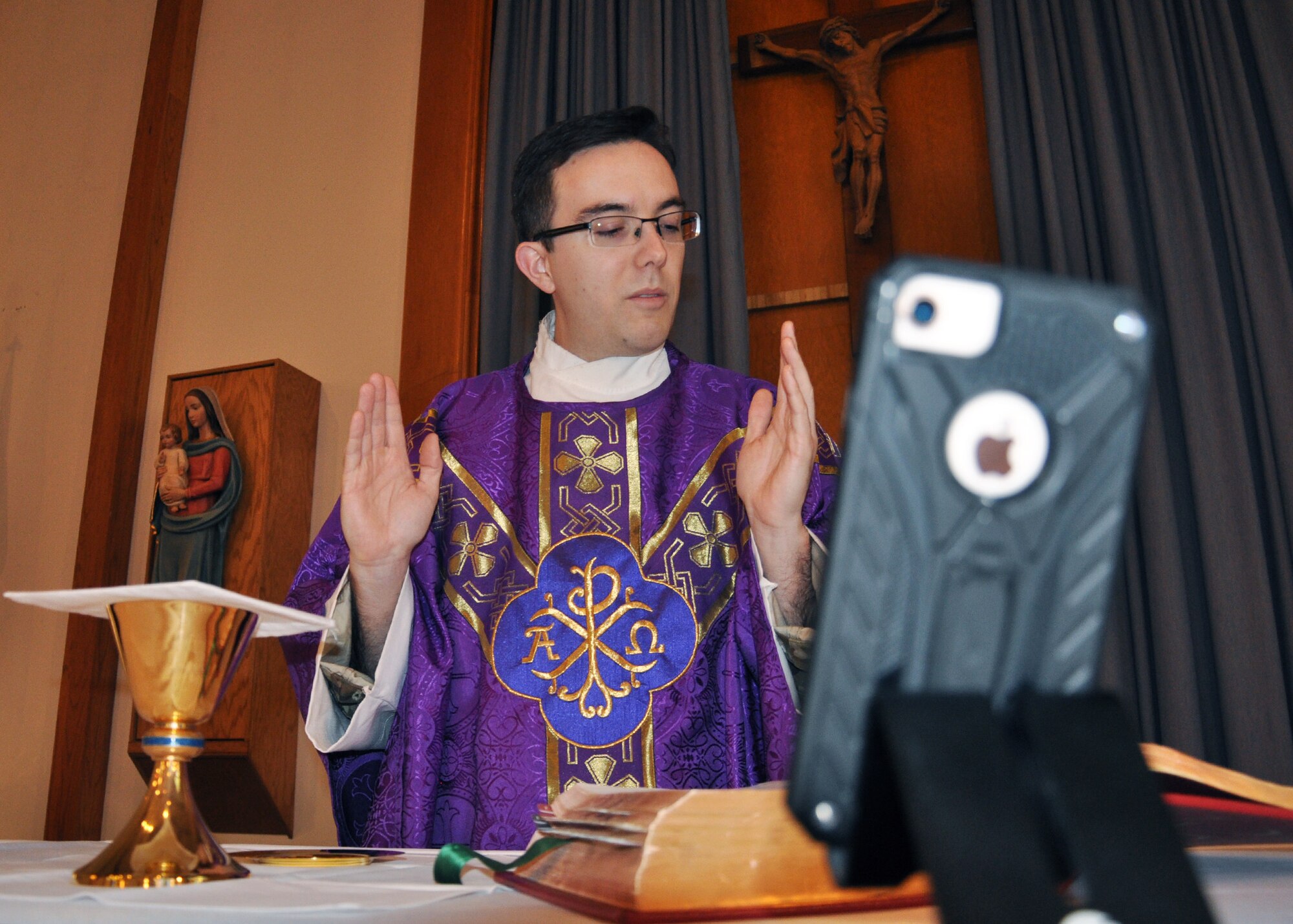 priest at alter preparing the gifts offered before communion during a virtual mass with a cell phone in foreground.