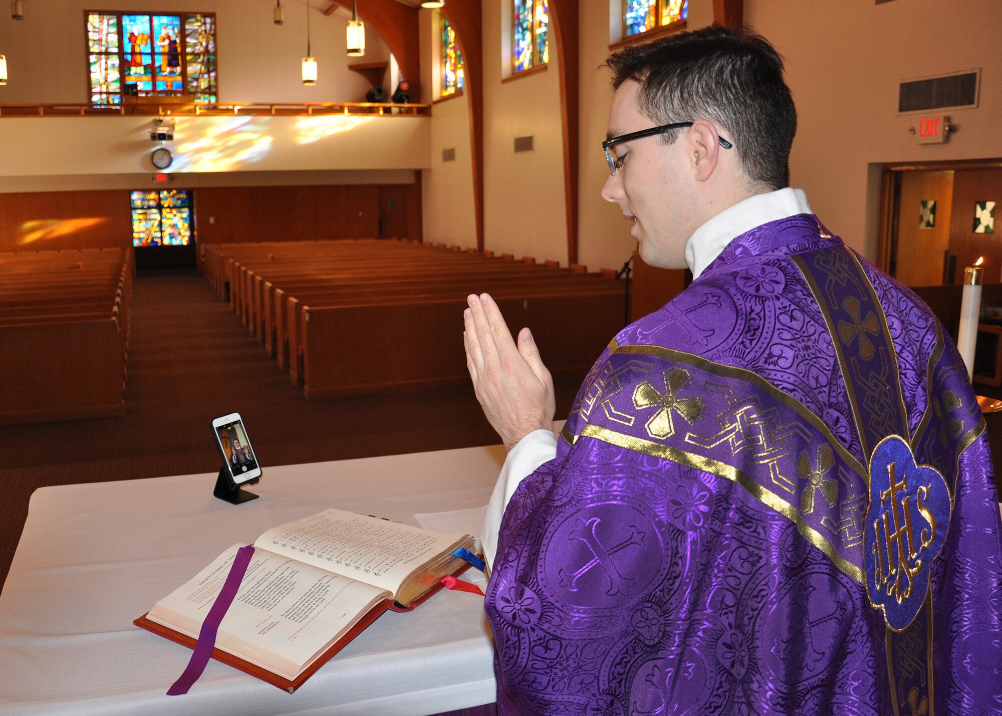 priest at altar reading the gospel, with empty pews in the background while recording virtual mass with a cell phone.