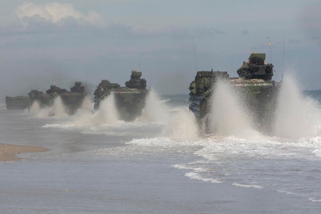 Marine Corps assault amphibian vehicles drive in a line through surf on a beach.