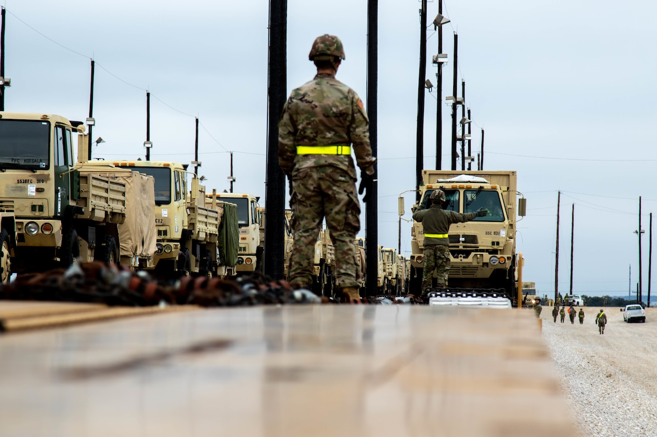 A military person guides the transport of military vehicles in a rail yard.