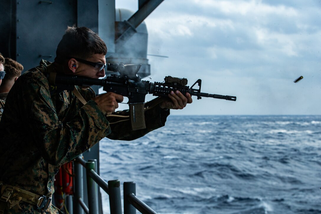 A Marine fires a service rifle over water from aboard a ship.