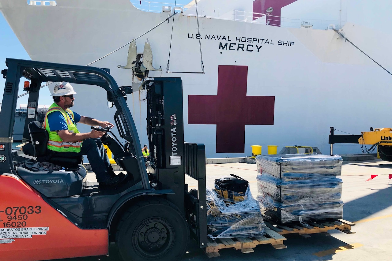 In the foreground, a forklift moves supplies. Behind the forklift is a Navy ship.