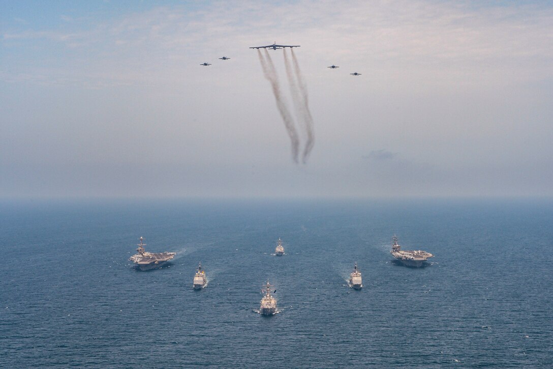 Ships moves through waters as aircraft fly above.