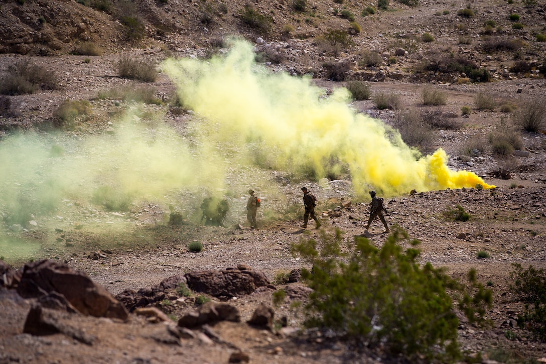 A group of Marines walk across a desert landscape while yellow smoke billows from behind them.