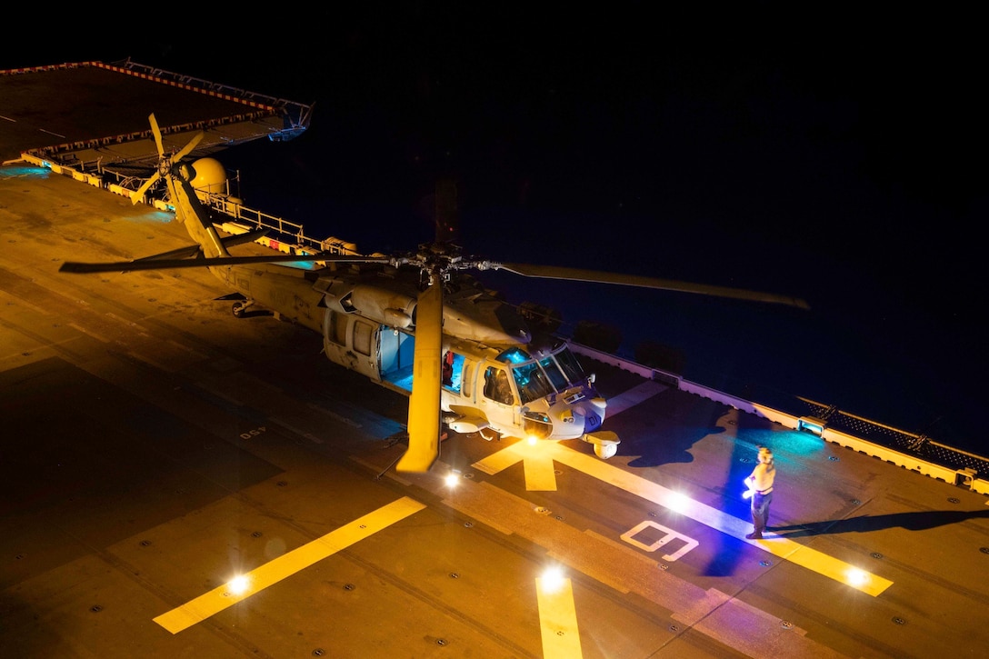 A sailor signals to a helicopter on the deck of a ship.