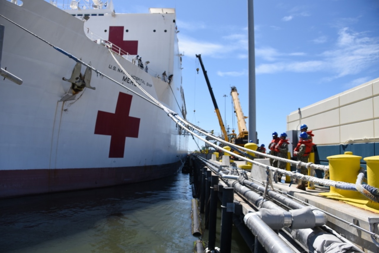 Sailors wearing blue helmets work beside a docked ship marked with red crosses.