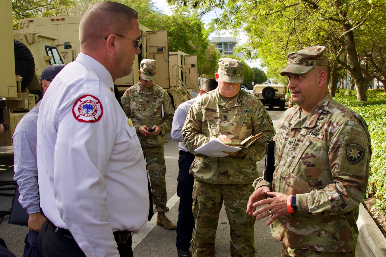 A soldier talks with a member of the fire department while standing outside near military vehicles.