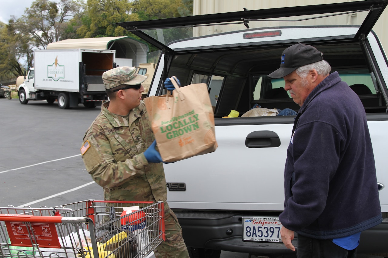 A soldier loads groceries into the back of a truck while an older man stands nearby.