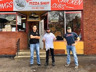 One man in black shirt with blue jeans and black shoes stands with crossed arms, next to man in white t-shirt and black pants with thumbs up, and a third man in blue shirt with red lettering and blue jeans stands with hands on hips in front of a brick building that has a pizza shop sign.