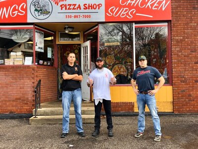 One man in black shirt with blue jeans and black shoes stands with crossed arms, next to man in white t-shirt and black pants with thumbs up, and a third man in blue shirt with red lettering and blue jeans stands with hands on hips in front of a brick building that has a pizza shop sign.