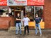 One man in black shirt with blue jeans and black shoes stands with crossed arms, next to man in white t-shirt and black pants with thumbs up, and a third man in blue shirt with red lettering and blue jeans stands with hands on hips in front of a brick building that has a pizza shop sign.