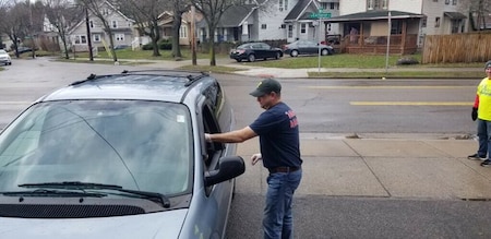 Man in blue shirt, blue jeans, and blue hat with white gloves on hands a bag to driver of grey van sitting in a parking lot.