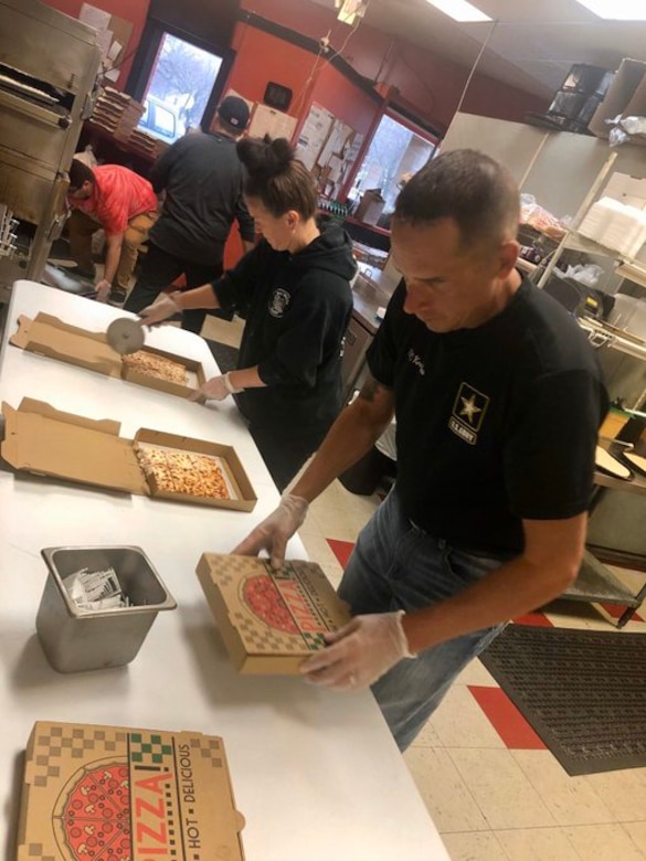 Man in black shirt, blue jeans and white gloves holds pizza box, while woman in black shirt cuts pizza in box.
