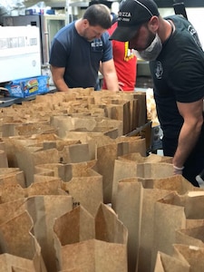 Man in black t-shirt and black hat and beard covered by guard checks brown bags while man in blue shirt and man in red shirt stands in background.