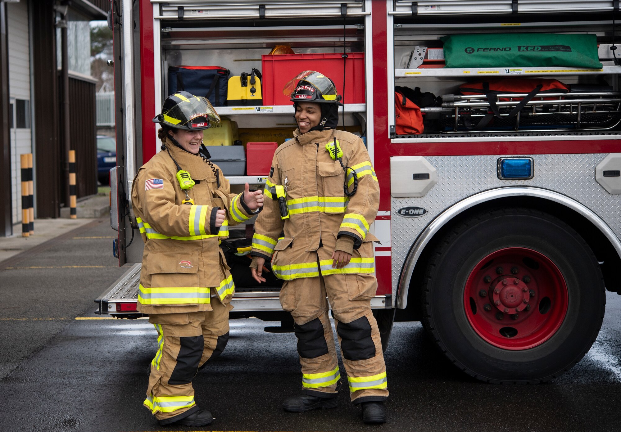 Two female firefighters laugh in front of a firetruck