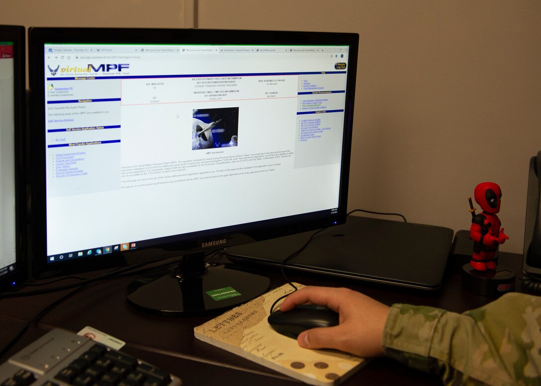 Image of an airman's hand holding mouse in the foreground with computer screen in background with the virtual military personnel flight website on computer screen