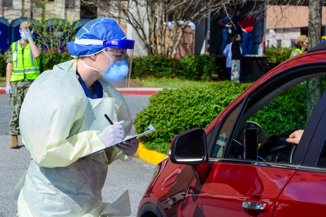 A sailor wearing a protective mask, gloves and smock leans in toward the driver's side of a car taking notes.